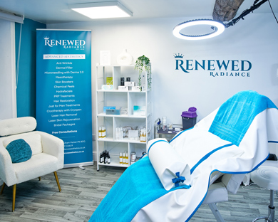 spa treatment room featuring a comfortable chair and a treatment bed with blue and white linens in front of a branded banner and shelving displaying beauty products and services for radiant skin